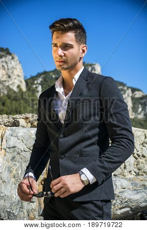 Young handsome man in classical suit on beach holding sunglasses while looking away. Sea waves on background
