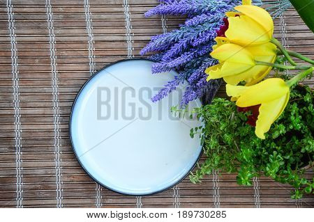 White ceramic plate with blue outer ring line.  Fresh green plants with exotic orchids of yellow and red flowers, and artificial lavender.  Vintage bamboo matt as background.