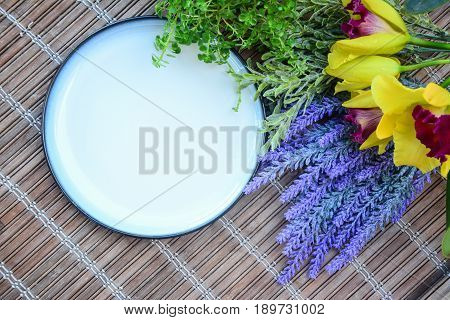 White ceramic plate with blue outer lines surrounded by fresh green plants, yellow and red color orchid and artificial lavender flower.  Vintage bamboo matt as background.