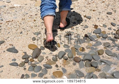 A man with jeans and black sandal walking along the sea pebbles beach.