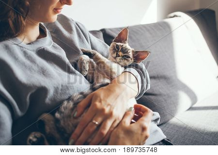 Happy young woman with her Devon Rex cat. Home pets. Young woman is holding and hugging her cute curious Devon Rex cat.