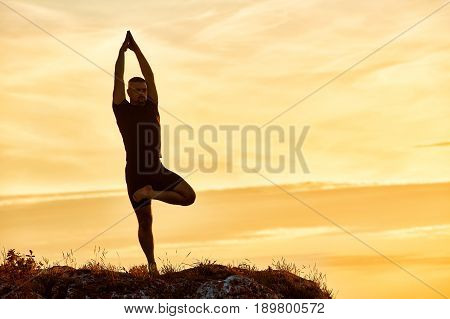 Silhouette of the man practicing yoga on the hill against beautiful sunset. Doing yoga exercises on the nature. Horizontal photo of young sportsman. Beautiful landscape and sky with clouds. Concept of the healthy lifestyle.