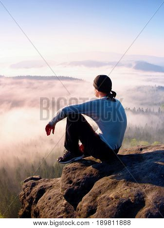 Man sit on the peak of rock and watching into colorful mist and fog in forest valley.