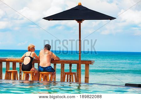 Successful retirement recreation summer vacation concept. Retired mature couple enjoying beautiful sunny day in swimming pool at beach club. Happy senior woman and man sitting at poolside beach bar.