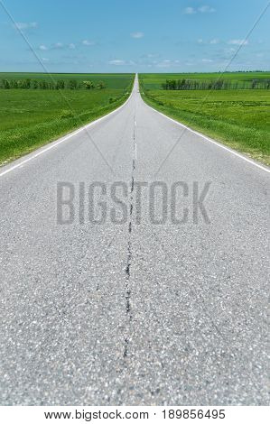 A wide asphalt road between green fields stretching into the blue sky