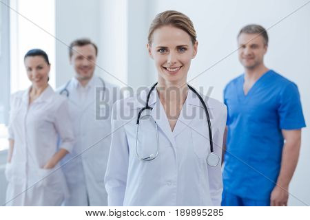 Visit our clinic for professional health checkup . Beautiful experienced mature medic standing in the clinic and smiling while her colleagues expressing happiness in the background