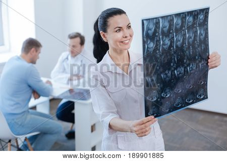 Helping more experienced colleague. Professional delighted smiling nurse working and analyzing mrt scan while doctor consulting patient in the background