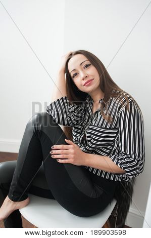 A beautiful brunette girl with long hair in a striped blouse and leather leggings sits on a white chair and smiles one hand is on her knees the second is holding her head.