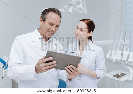 What can you advice me. Handsome smiling doctor holding tablet in both hands looking downwards while giving examples to his intern