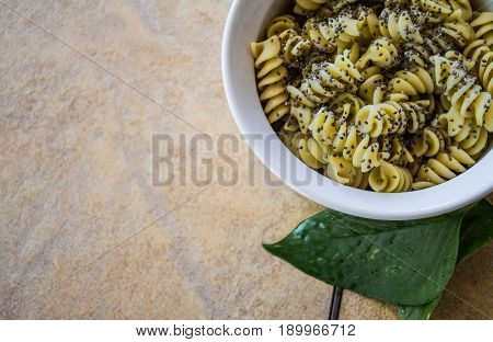 Rotini pasta swirls topped with black chia seeds in a white bowl on brown stone table. Copyspace on left side of food items.