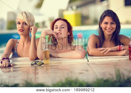 Young attractive blonde, brunette and red haired female bathers posing on edge of swimming pool at summer