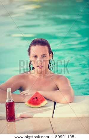 Portrait of young female bather in swimming pool with slice of watermelon and beverage by railing pool