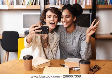 Two cheerful young girls students studying at library, taking a selfie