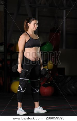 Pregnant female athlete doing dumbbell power snatch overhead press workout in dark gym