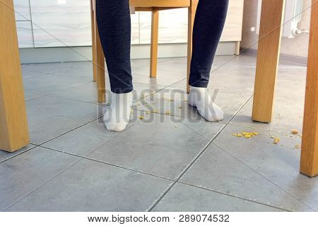 Woman Eats At The Table And Crumbles Corn Flakes On The Floor. Legs And Floor, Front View.