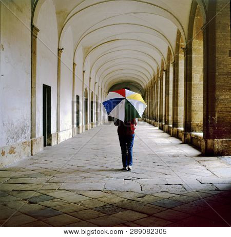 Lady With Multi Colored Umbrella Walking In The Galleries Of The Central Square Of Marciac, France. 