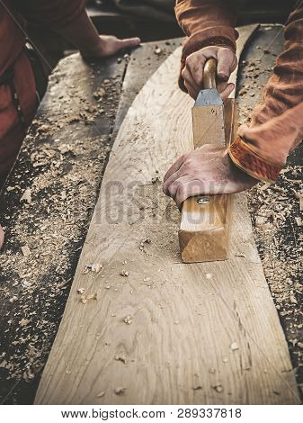 Carpenter In Medieval Cotton Clothes Working With A Wood By Plane. Man Manually Hews A Wooden Board 