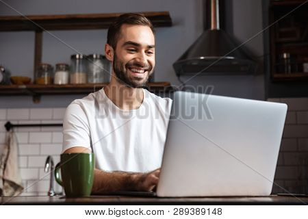 Photo of unshaved man 30s wearing casual t-shirt using silver laptop while sitting at table in modern apartment