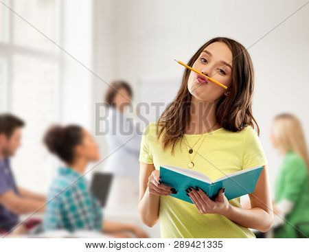 high school, education and people concept - foolish teenage student girl in yellow t-shirt with diary or notebook and pencil-mustache over classroom and teacher background