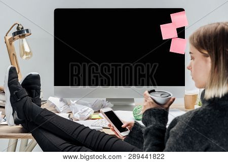 Selective Focus Of Girl Sitting At Messy Workplace With Thermomug And Using Smartphone Isolated On G