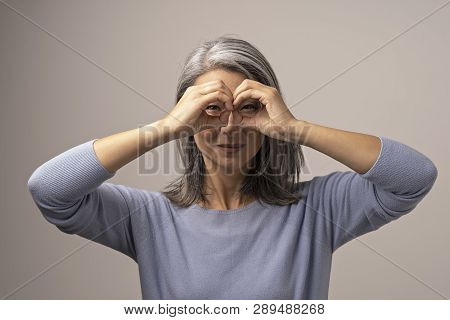 Beautiful Grey-haired Woman Squints Her Eyes While Looking Through Her Hands As If They Are Glasses.