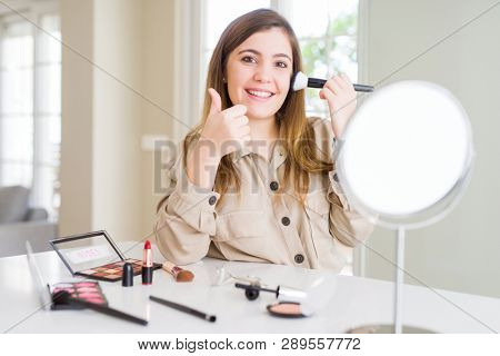 Beautiful young woman using make up cosmetics applying color using brush happy with big smile doing ok sign, thumb up with fingers, excellent sign