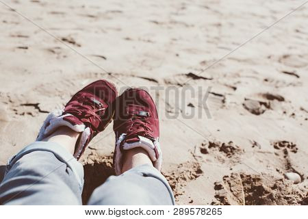 Feet Of Young Woman In Sport Footwear And Jeans Relaxing After Hiking On Sand Beach. Pov. Adventure 