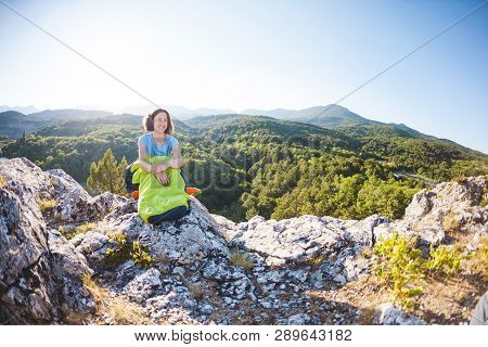 A Woman Is Sitting In A Sleeping Bag At Sunset. Camping On Top Of A Mountain. A Smiling Girl Rests A