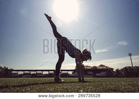 Young Slim Blond Woman At Green Grass Of Stadium In The Morning Making Exercises. Girl Do One-legged
