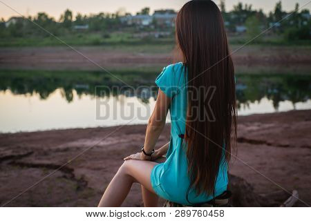 Young Girl In Turquoise Dress Sitting On A Stump On The Shore