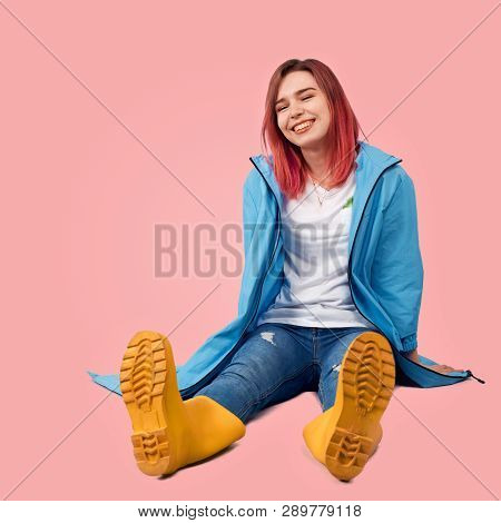 Cheerful Girl Sitting On The Floor. She Wearing A Blue Coat And Yellow Rubber Boots. Joyful Laughter