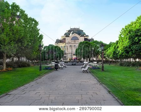 Kosice, Slovakia - May 2, 2018: Green spring park from the back of the Kosice State Theatre. Young grass, flowers, vintage street lamps and benches along alley against backdrop of theater building