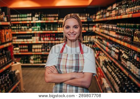 Young Woman Stand In Grocery Store Among Alcohol Shelfs. She Hold Hands Crossed And Smile. Posing On