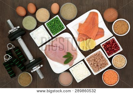 Body building dumbbell weights and hand grippers with high protein lean meat, fish and dairy, dietary supplement powders, vegetables, fruit and grains. Top view on oak wood table.