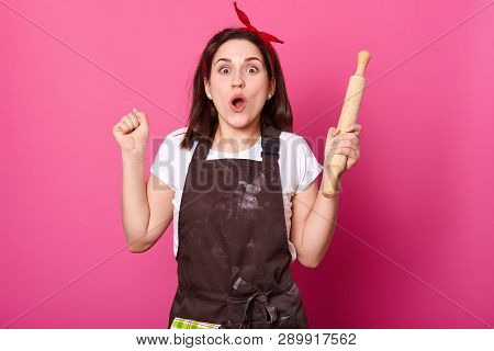 Young Baker Against Pink Wall Looks At Camera, Bends Elbows, Holds Rolling Pin. Charming Lady With H