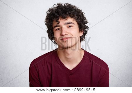 Close Up Shot Of Curly Haired European Teenager With Little Beard, Wears Maroon Shirt. Young Smiling