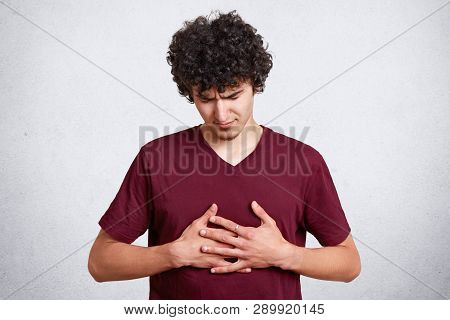 Portrait Of Upset Unhappy Teenager Dresses Maroon T Shirt, Keeps Hands On Chest Suffering From Pain 