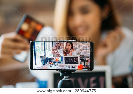 Happy Young Attractive Asian Ethnic Model Filming Vlog On Mobile Phone On Tripod And Showing Eyeshad