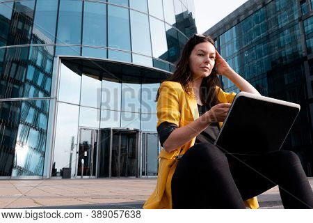 Freelancer With A Laptop Sat On The Steps Near The Business Center, A Young Woman In A Yellow Jacket