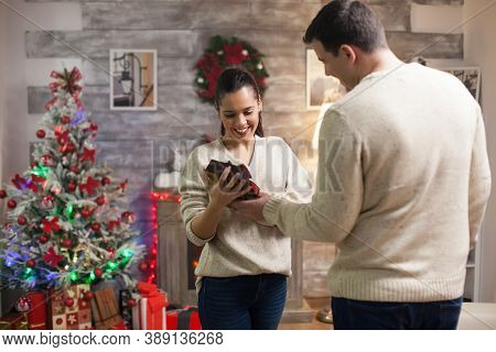 Tall Young Man Giving His Girlfriend A Gift On Christmas Day. Happy Wife And Xmas Tree.