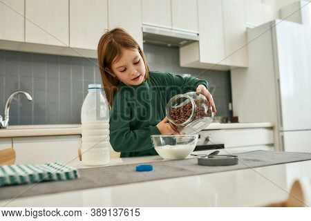 A Small Cute Girl Pouring Cereal Balls Into A Bowl With Milk For Breakfast Stainding In A Large Kitc