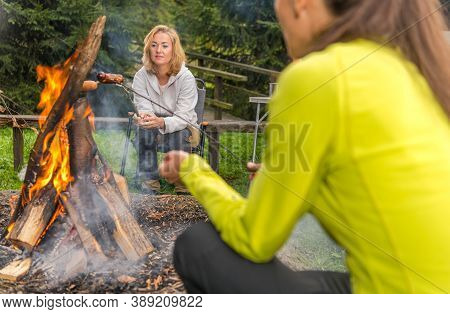 Caucasian Female Friends In Their 40s Hanging In Front Of Campfire Grilling Sausages And Having Fun.