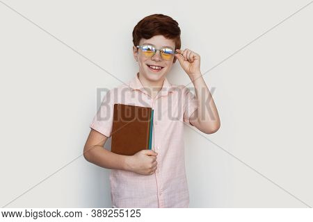 Caucasian Ginger Boy With Glasses Is Smiling At Camera Holding Some Books On A White Studio Wall In 