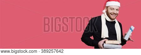 Bearded Caucasian Man Is Showing At Camera Some Presents Smiling On A Red Wall With Free Space In Bl