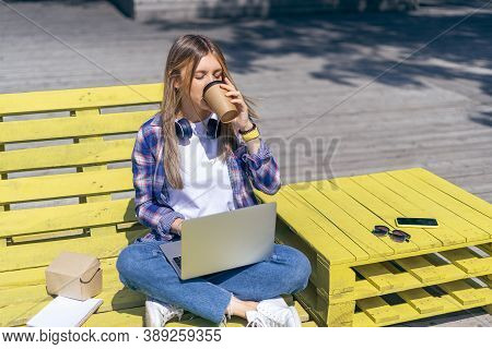 Beautiful Woman Working At Notebook Sit Down On Bench Outside And Drinking Coffee. Using Computer An