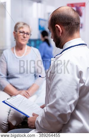 Doctor Holding Medical Document While Speaking With Senior Old Woman In Clinic Cabinet. Converstatio