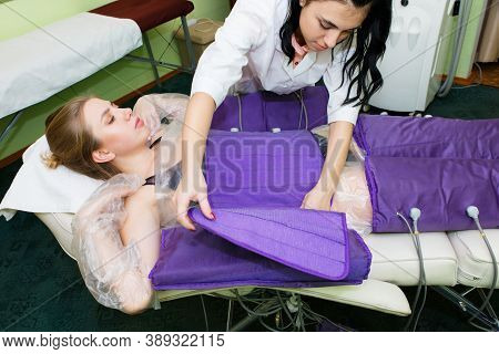 A Beautician With Dark Hair Prepares A Young Woman For The Pressotherapy Process. Wearing A Special 