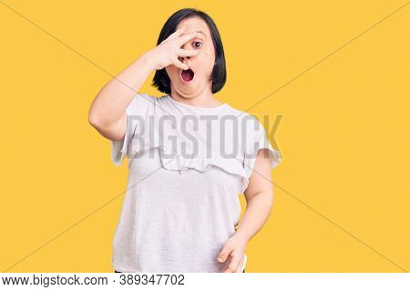 Brunette woman with down syndrome wearing casual white tshirt peeking in shock covering face and eyes with hand, looking through fingers with embarrassed expression. 