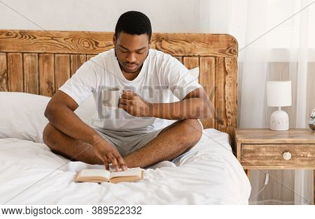 Morning Read. African American Man Reading Longread Books Drinking Coffee Sitting In Cozy Bed In Bed
