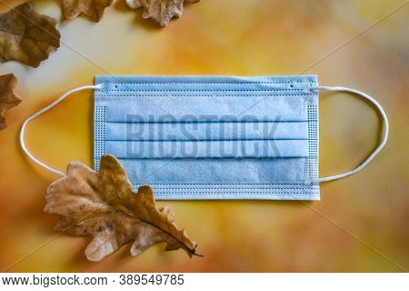 A Medical Protective Mask And Yellow Dry Oak Leaves Lie On A Bright Autumn Background. The Concept O